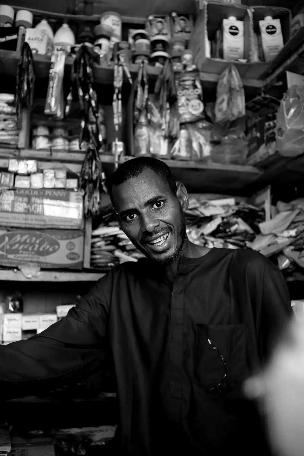 Un commerçant souriant dans sa boutique, devant des étagères chargées de produits.