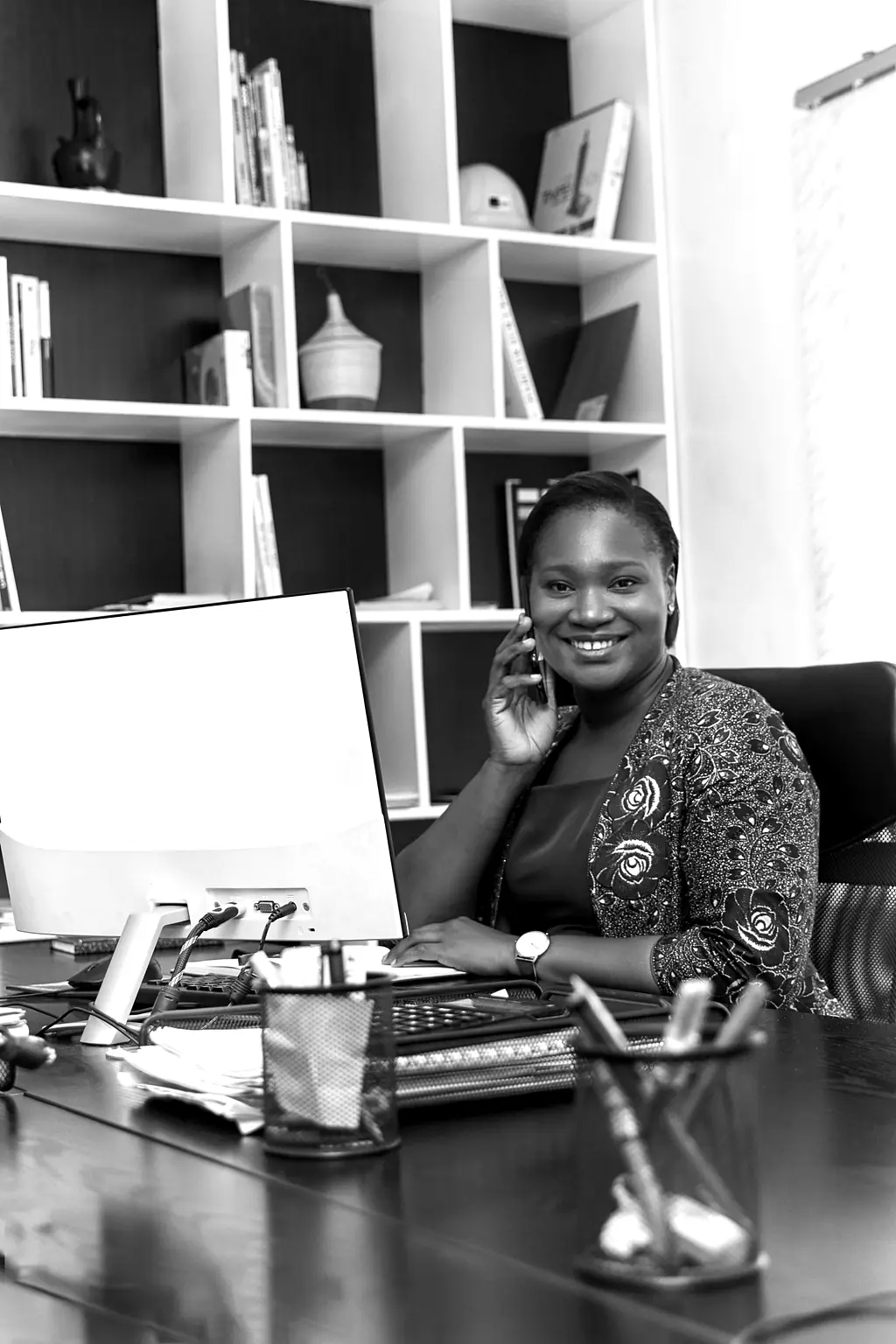 Une femme souriante, à son bureau, parle au téléphone devant un ordinateur.