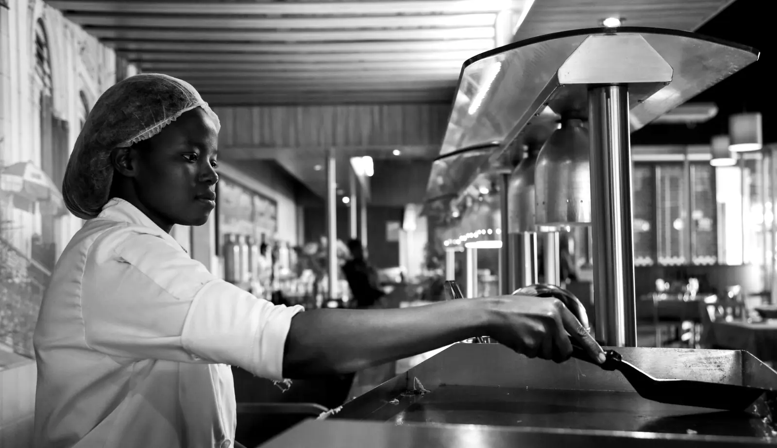 Une cuisinière en tenue blanche et charlotte travaille au passe d'une cuisine de restaurant.